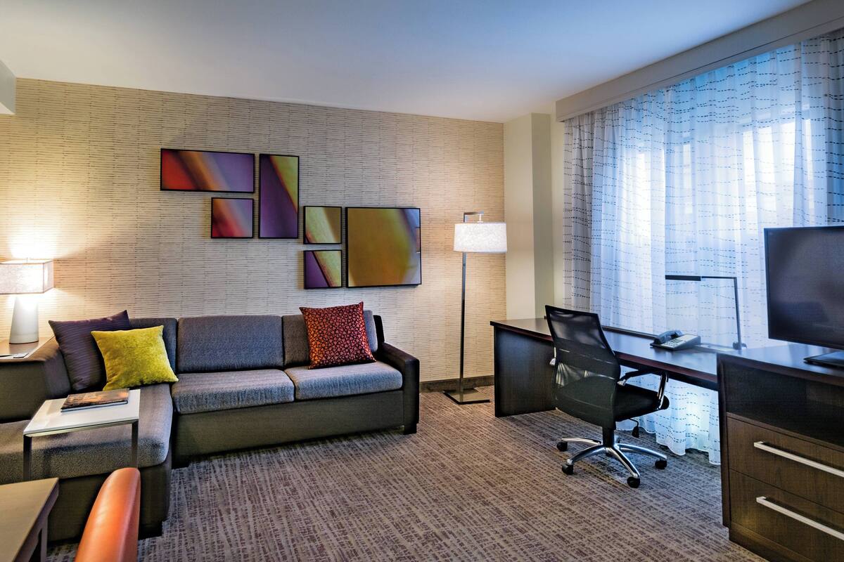 A modern hotel room with a sofa, desk, and chair, a flat-screen TV, and artwork on the wall.