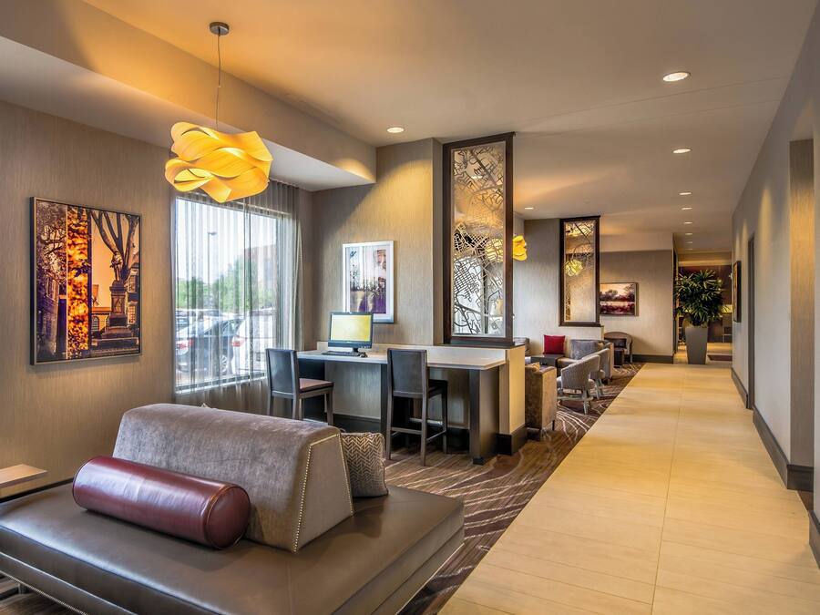 A modern hotel room with a sofa, desk, and computer. There are large windows, artwork on the walls, and a hallway with a plant.