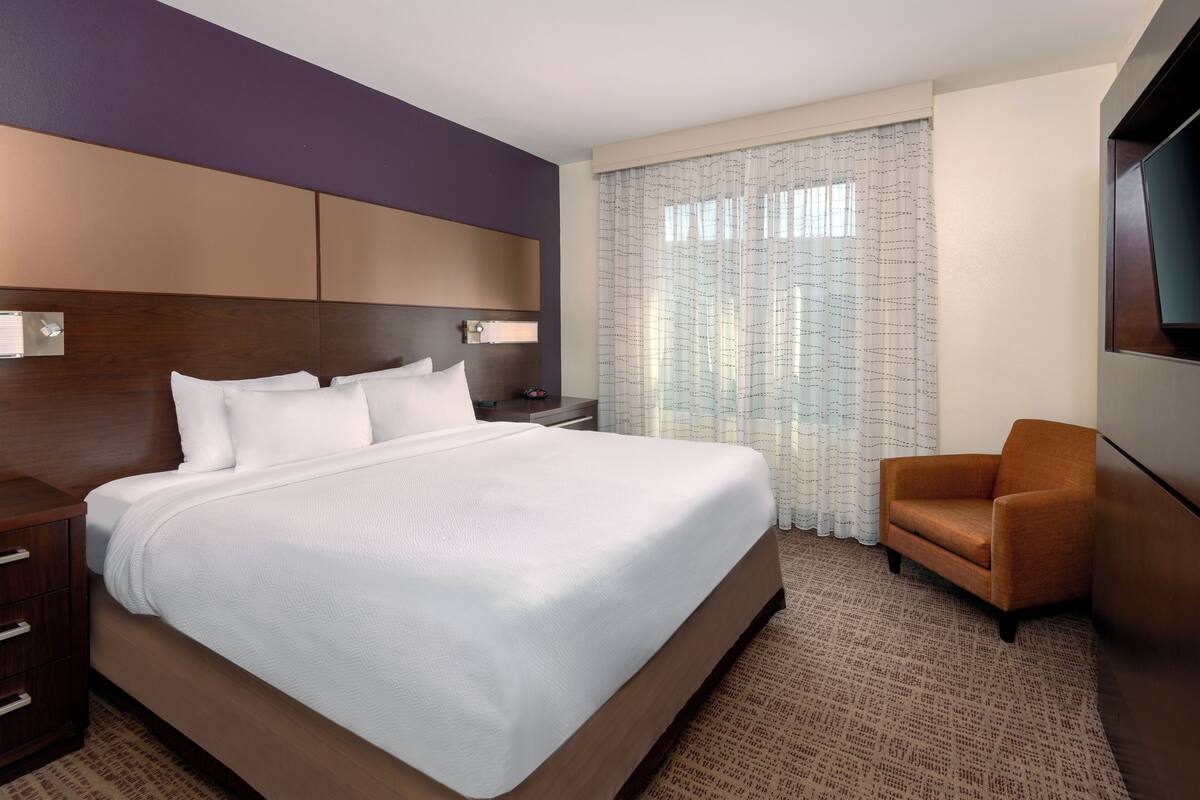 A hotel room with a large bed, a wooden headboard, a nightstand, a chair, and a TV mounted on the wall.