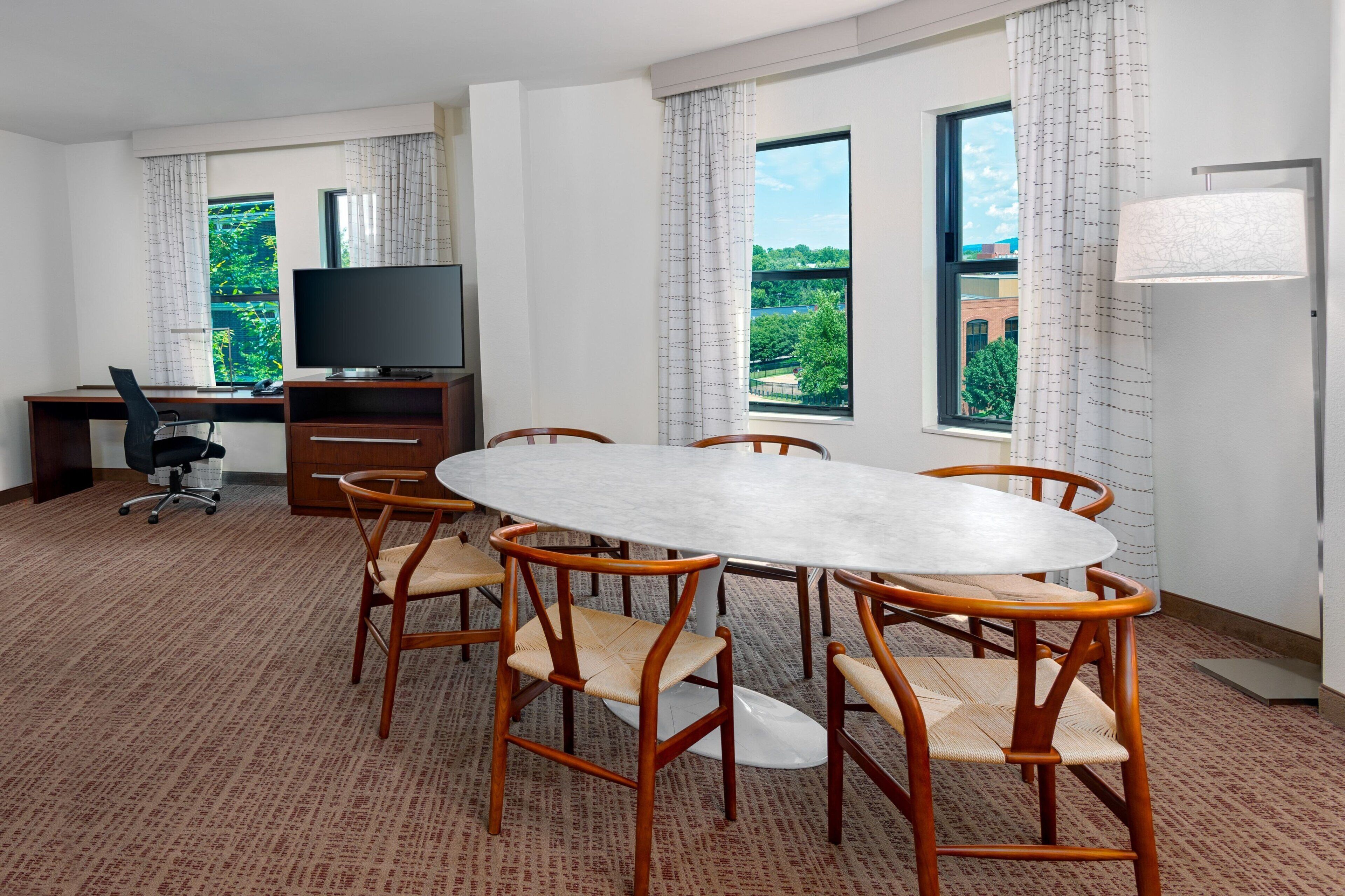 A conference room with a round table, six wooden chairs, a television on a desk, and large windows with curtains.