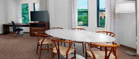 A conference room with a round table, six wooden chairs, a television on a desk, and large windows with curtains.