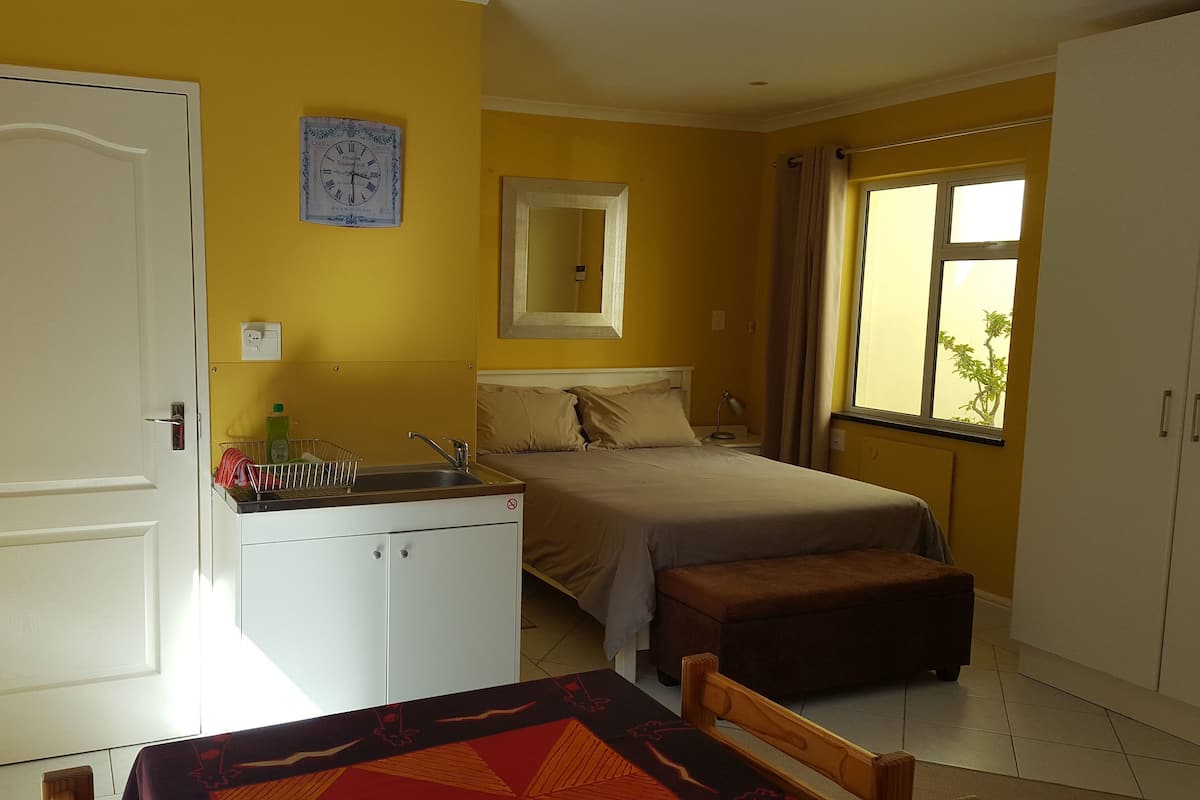 A compact hotel room with a bed, a small kitchenette, a window with curtains, and a door.