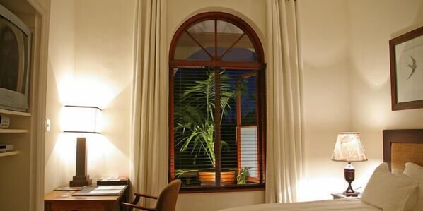 Una habitación de hotel con una cama, un escritorio con lámpara, una silla, una ventana con cortinas y un reloj fijado en la pared.