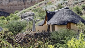 A thatched-roof house with a stone wall and a view of a cliff. - Vingerklip Lodge (Khorixas)
