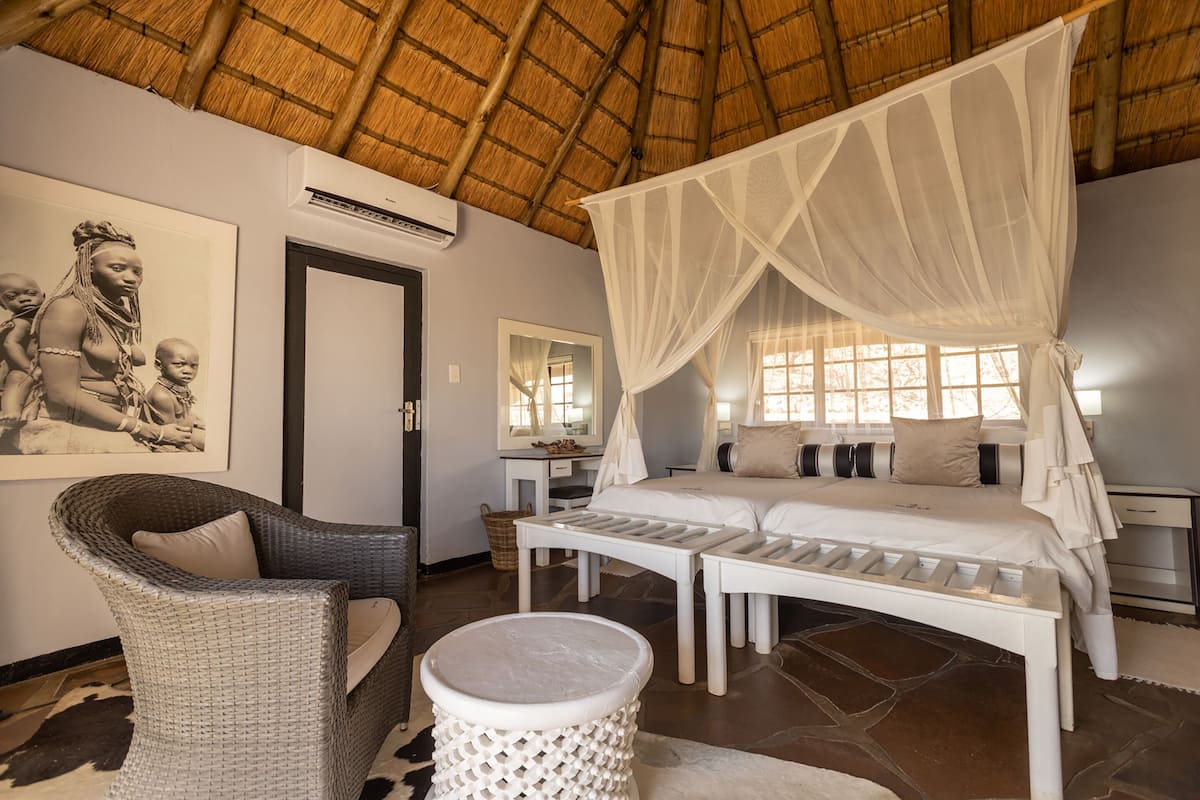 A bedroom with a bed, a wicker chair, a round stool, a mirror, and a wall-mounted air conditioner.