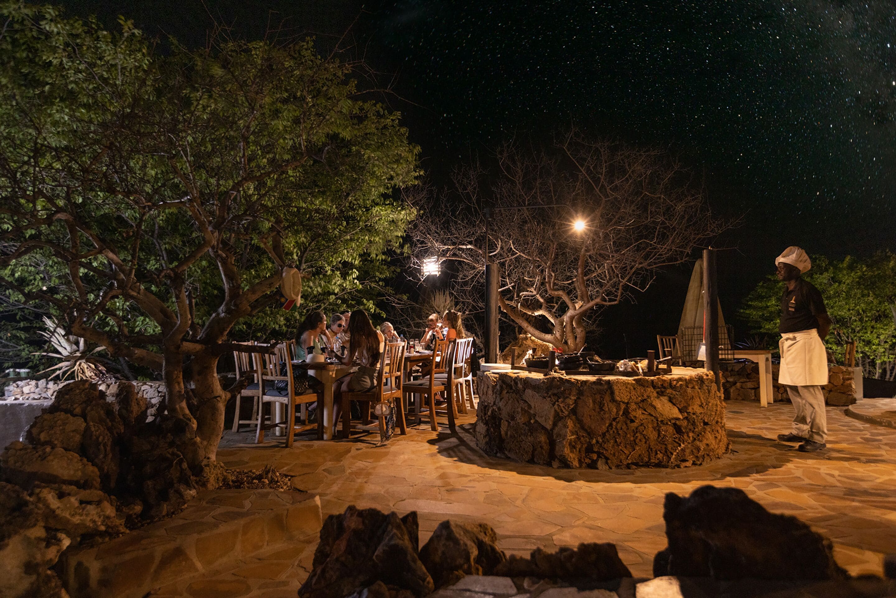 Un espacio de comedor al aire libre con mesas y sillas de piedra, rodeado de árboles y un muro de piedra.