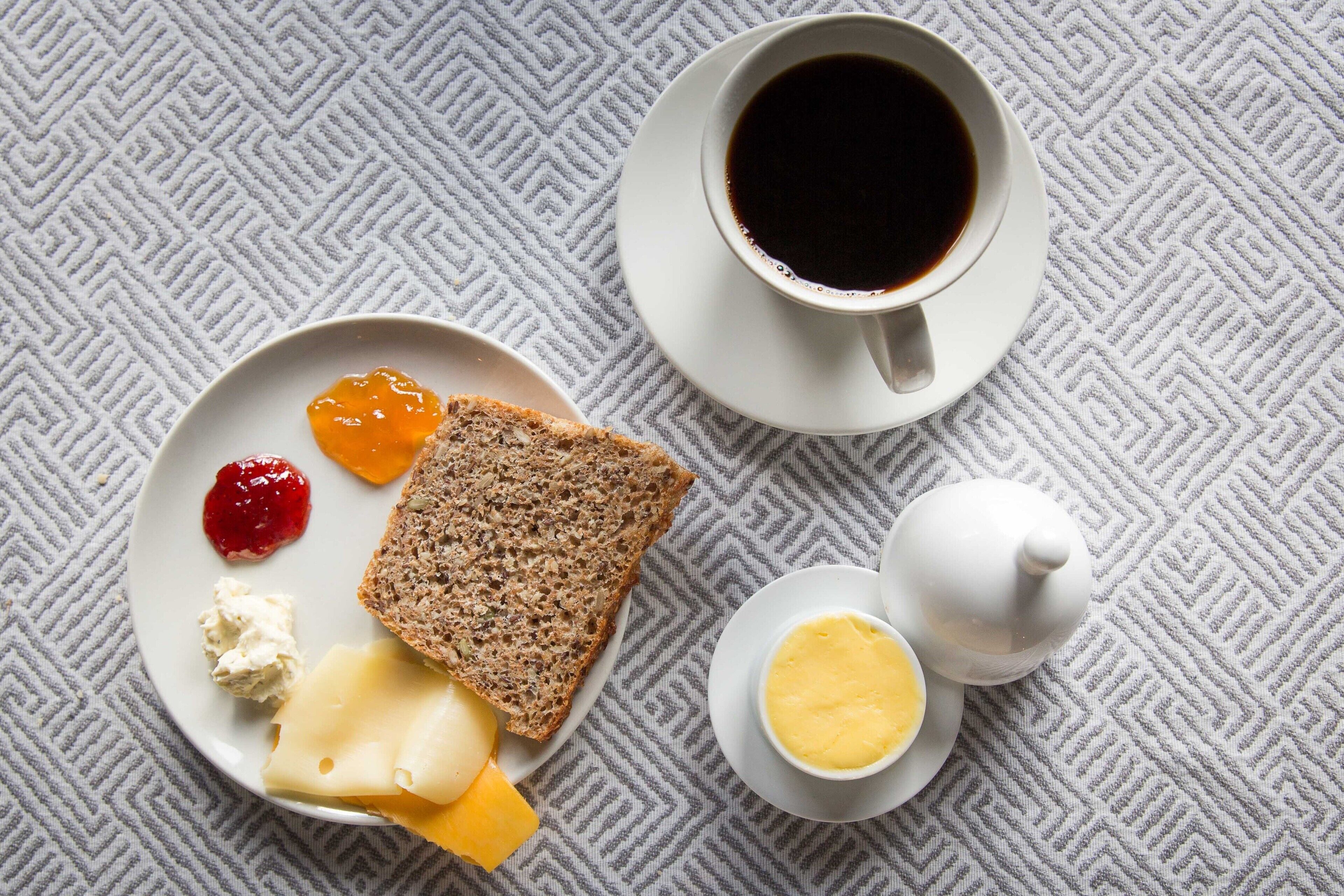 Ein Teller mit Käse, Marmelade, Butter und einer Scheibe Brot, eine Kaffeetasse und ein Teekessel auf einer gemusterten Tischdecke.