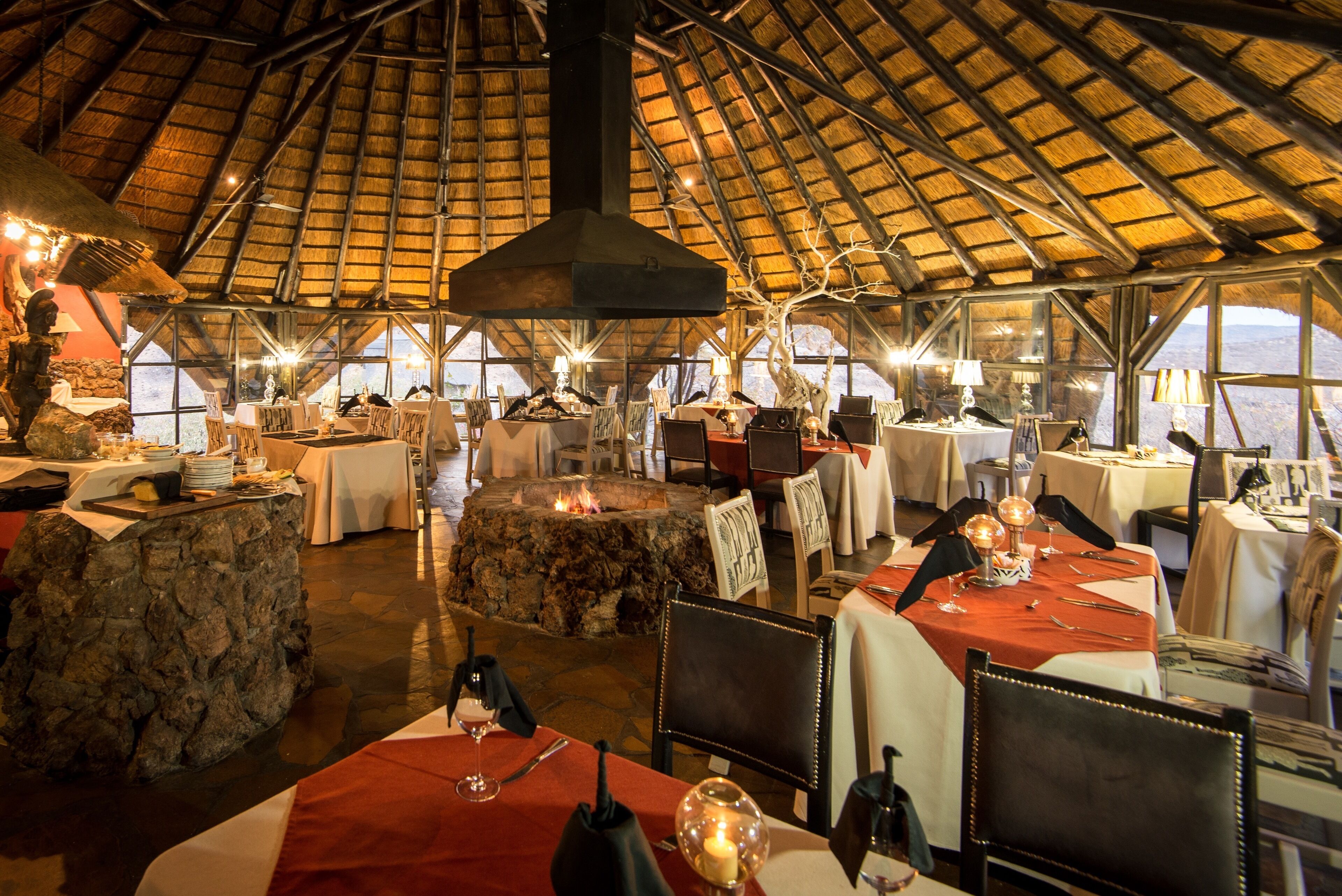 Un restaurant doté de tables en pierre et d’une cheminée en pierre, de grandes fenêtres et d’un toit de chaume élevé.