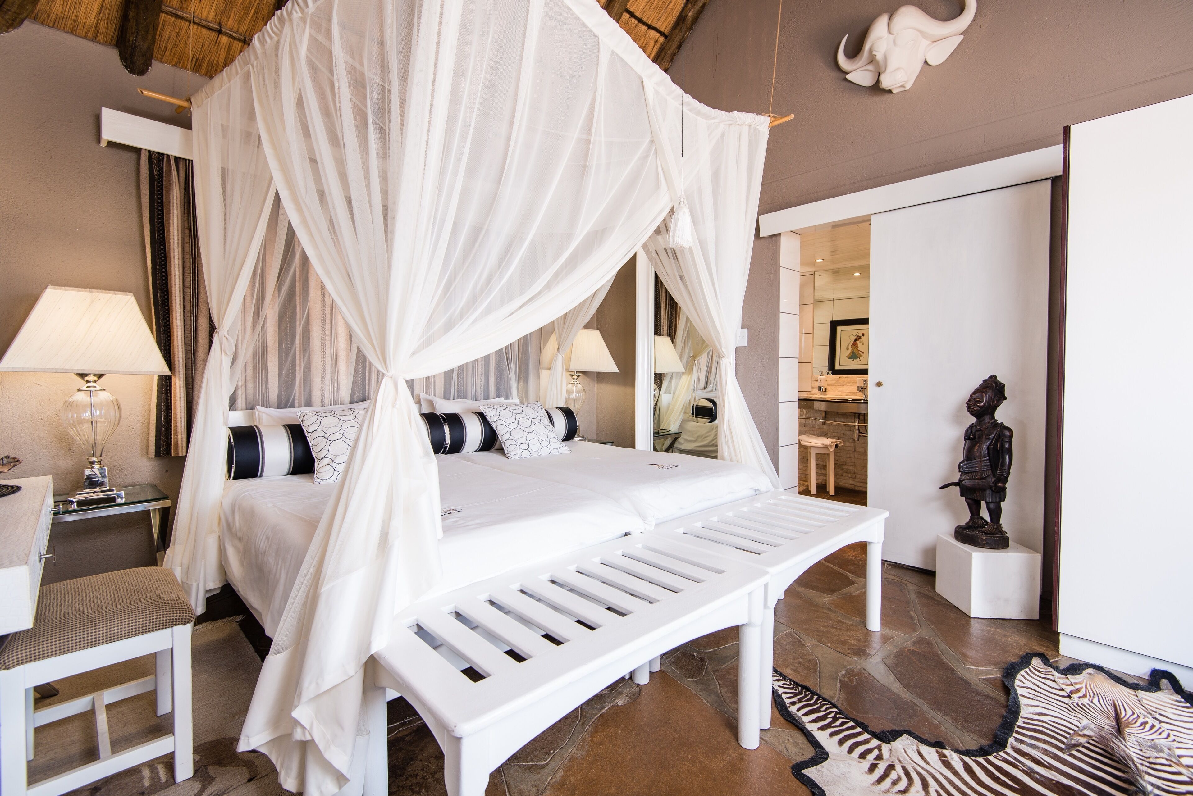 A bedroom with a canopy bed, a bedside table with a lamp, a bench, and a zebra-patterned rug.