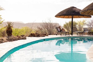 A pool area with lounge chairs and umbrellas, surrounded by desert vegetation and rocks. - Vingerklip Lodge (Khorixas)