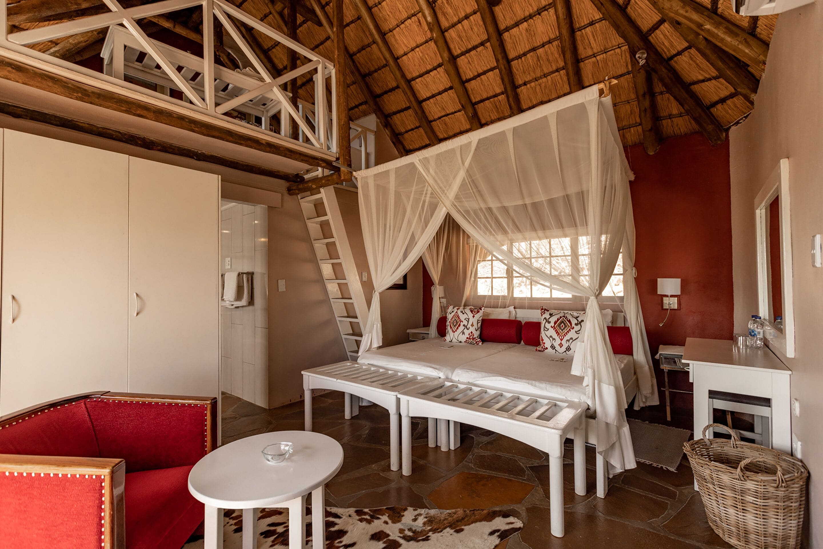 A bedroom with a canopy bed, a red chair, a white table, and a stone floor.