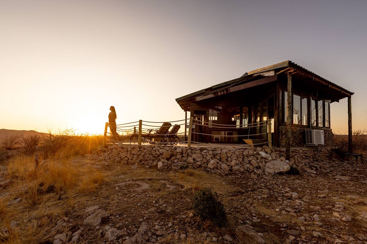 A rustic building with a stone foundation and a wooden structure, featuring a balcony with chairs and a person standing nearby.