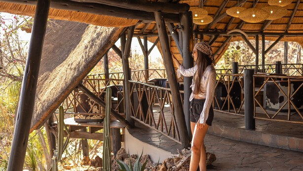 A covered outdoor area with a wooden deck, stone flooring, and a metal railing. - Vingerklip Lodge (Khorixas)
