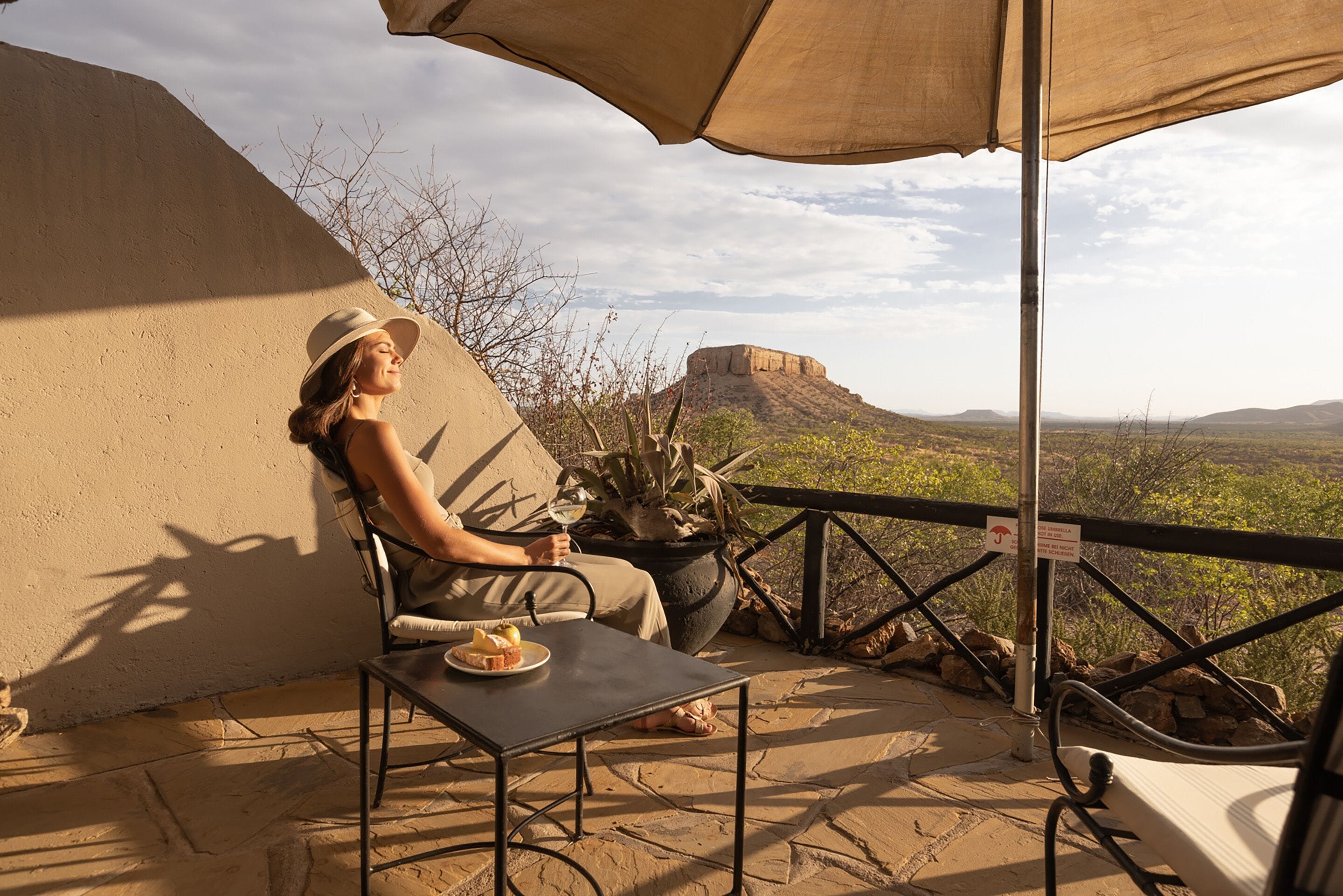 A person seated at a table with a plate of food, under a large umbrella, overlooking a scenic landscape with a prominent hill in the background.