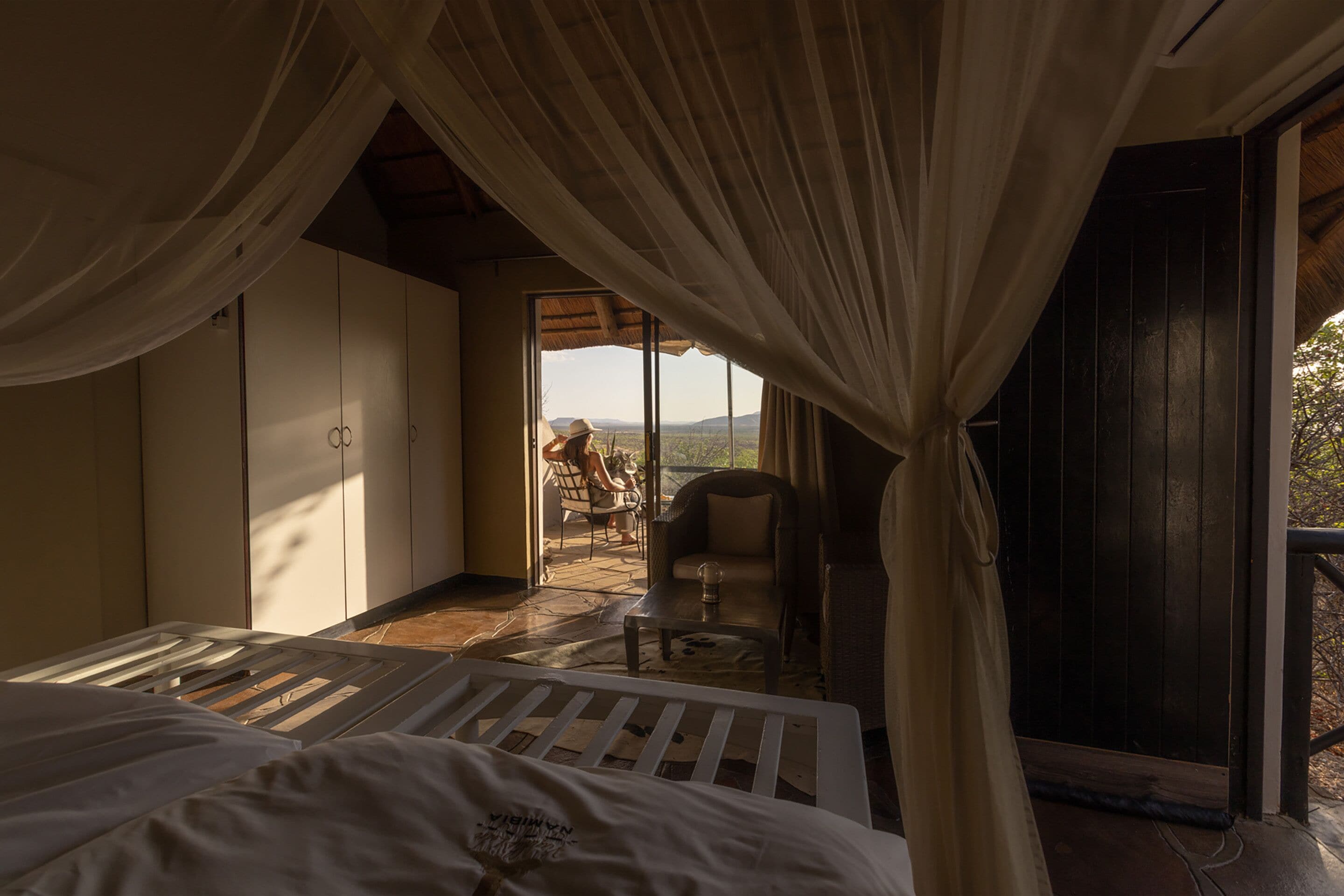 A bedroom with a large bed, a view of a balcony with a chair, and a person sitting outside.