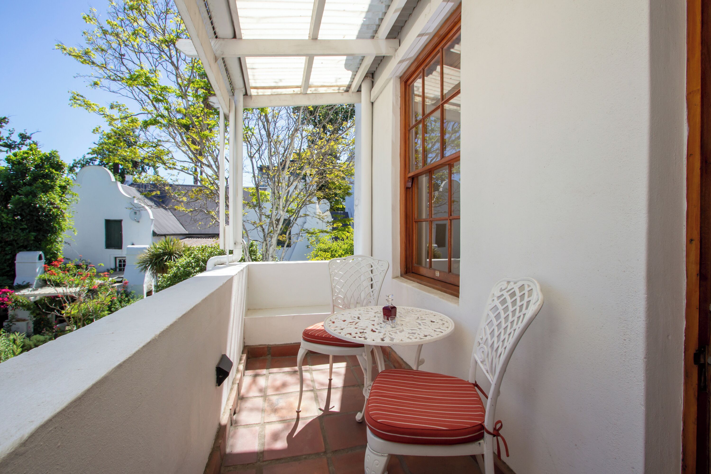 Un balcón con sillas y una mesa blancos, una ventana y vistas a zonas verdes y edificios.
