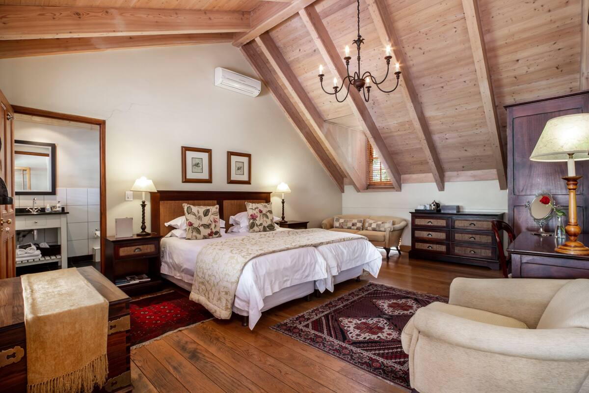 A bedroom with a sloped ceiling, wooden beams, a large bed with floral bedding, bedside lamps, a dresser, and a mirror.