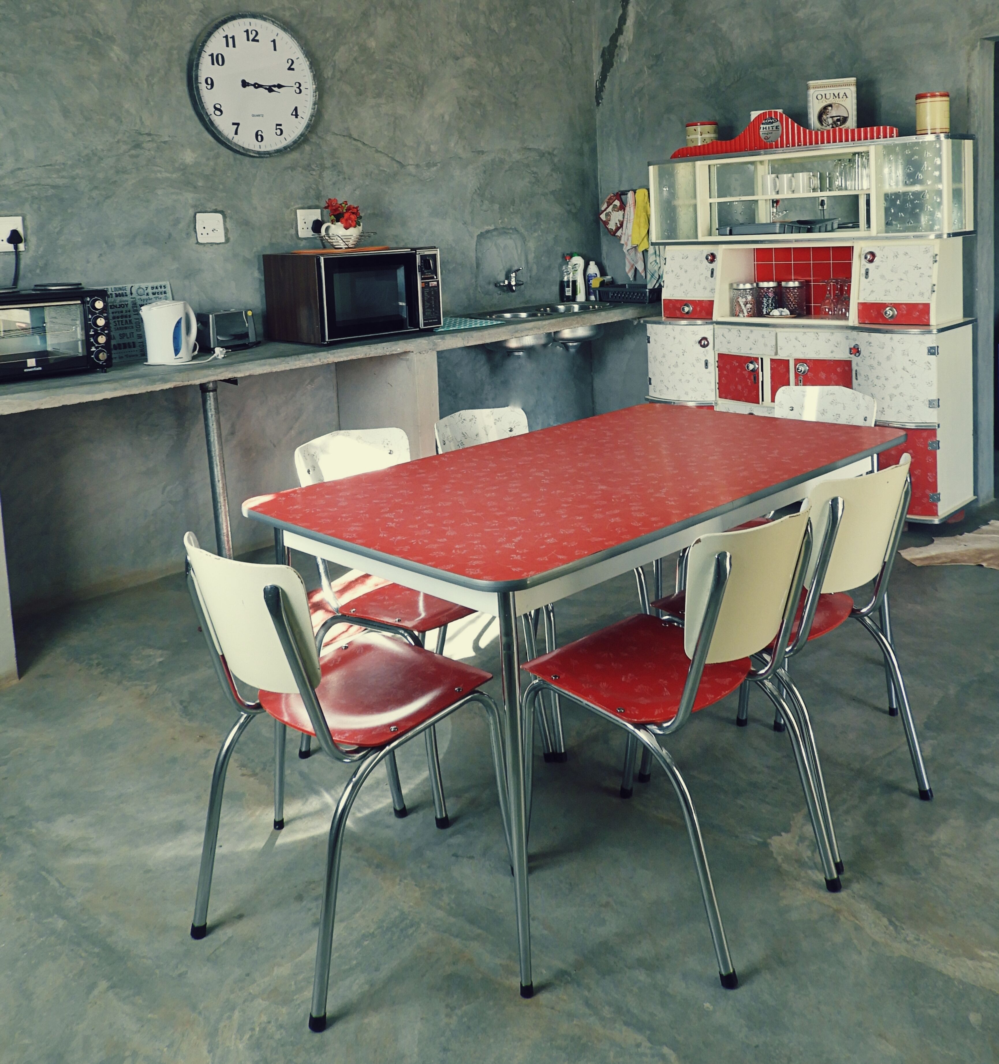 A kitchen with a red table and four chairs, a clock on the wall, and vintage appliances.