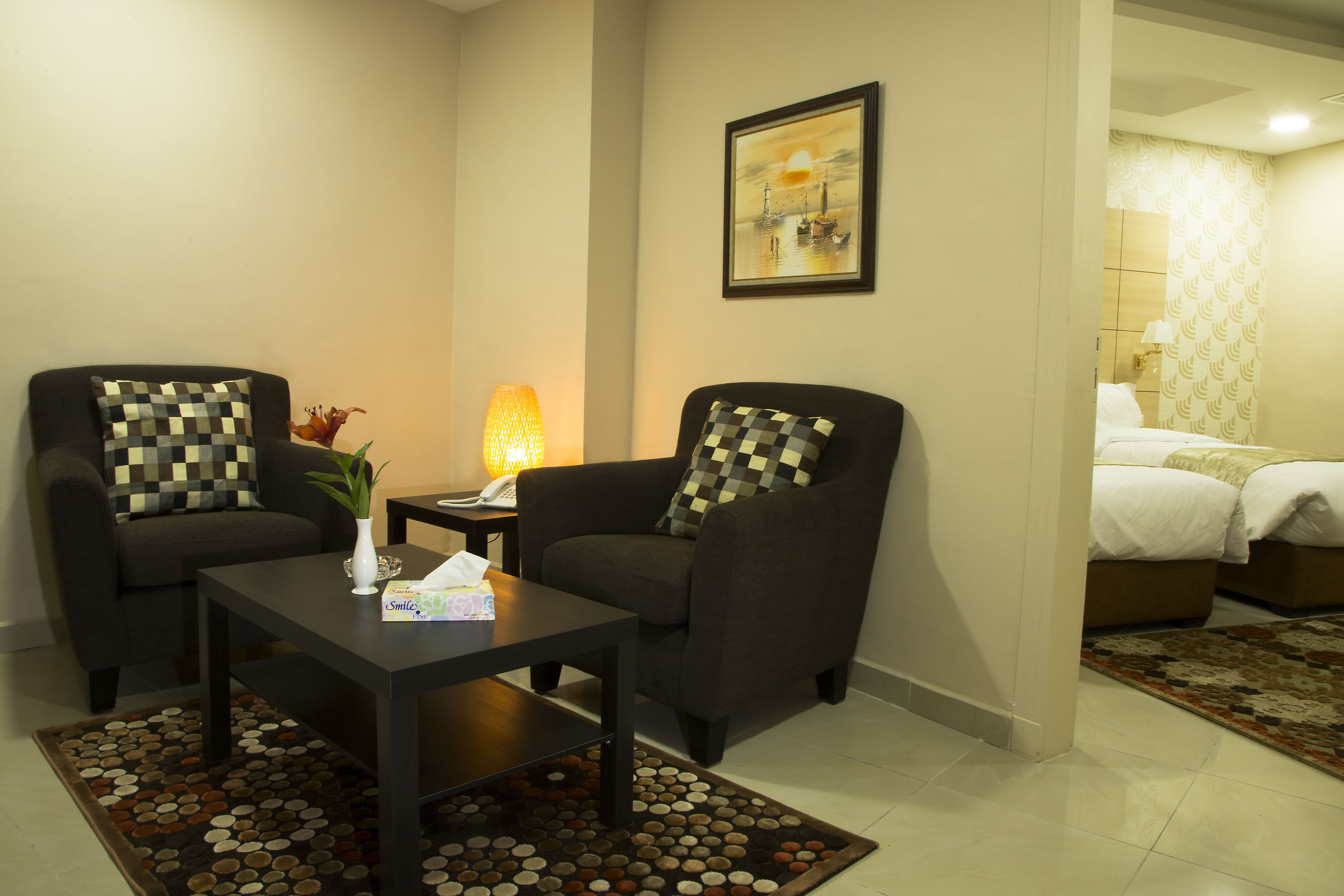Een hotelkamer met twee fauteuils, een bijzettafeltje, een lamp, een schilderij en een bed met een beddeken met dessin.