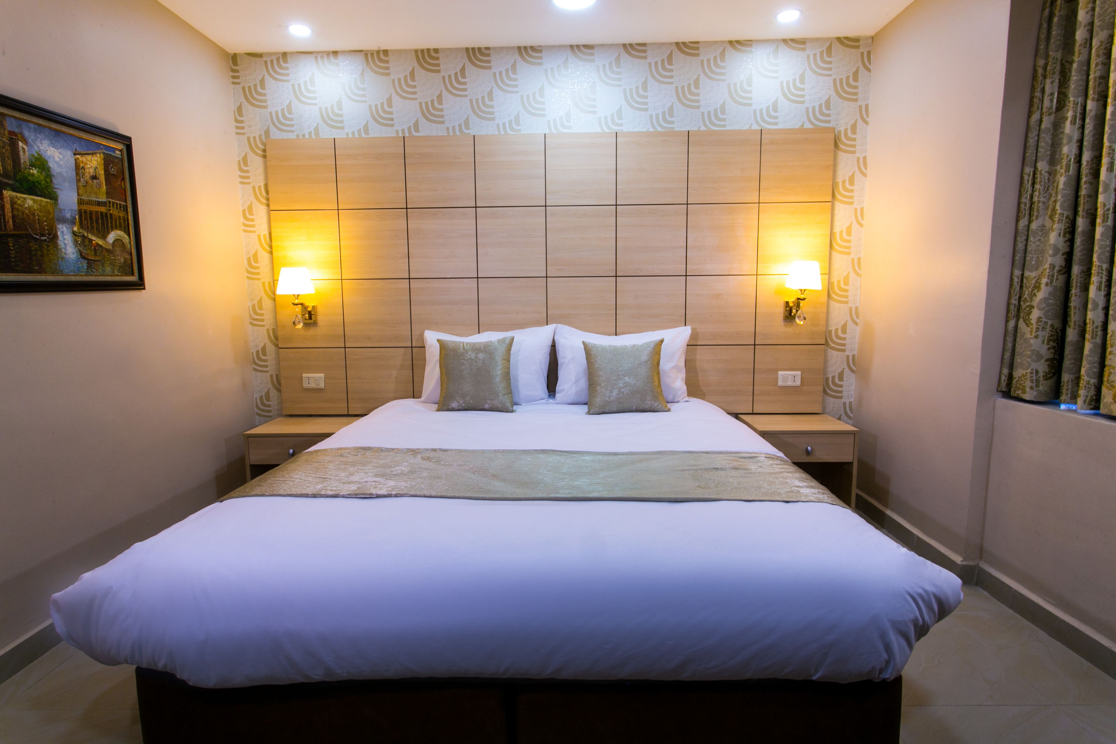A neatly made bed with a headboard, two bedside lamps, and a painting on the wall.