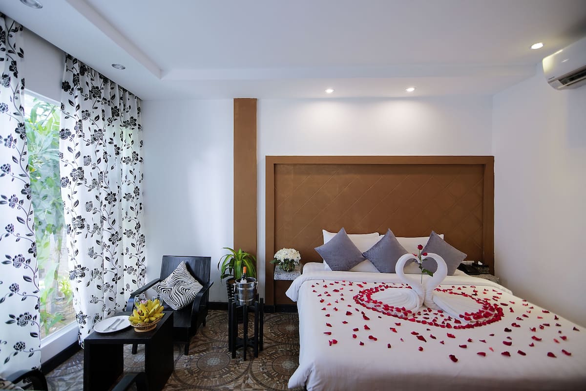 Habitación de hotel con cama, escritorio, silla, planta en maceta y ventana con cortinas florales.
