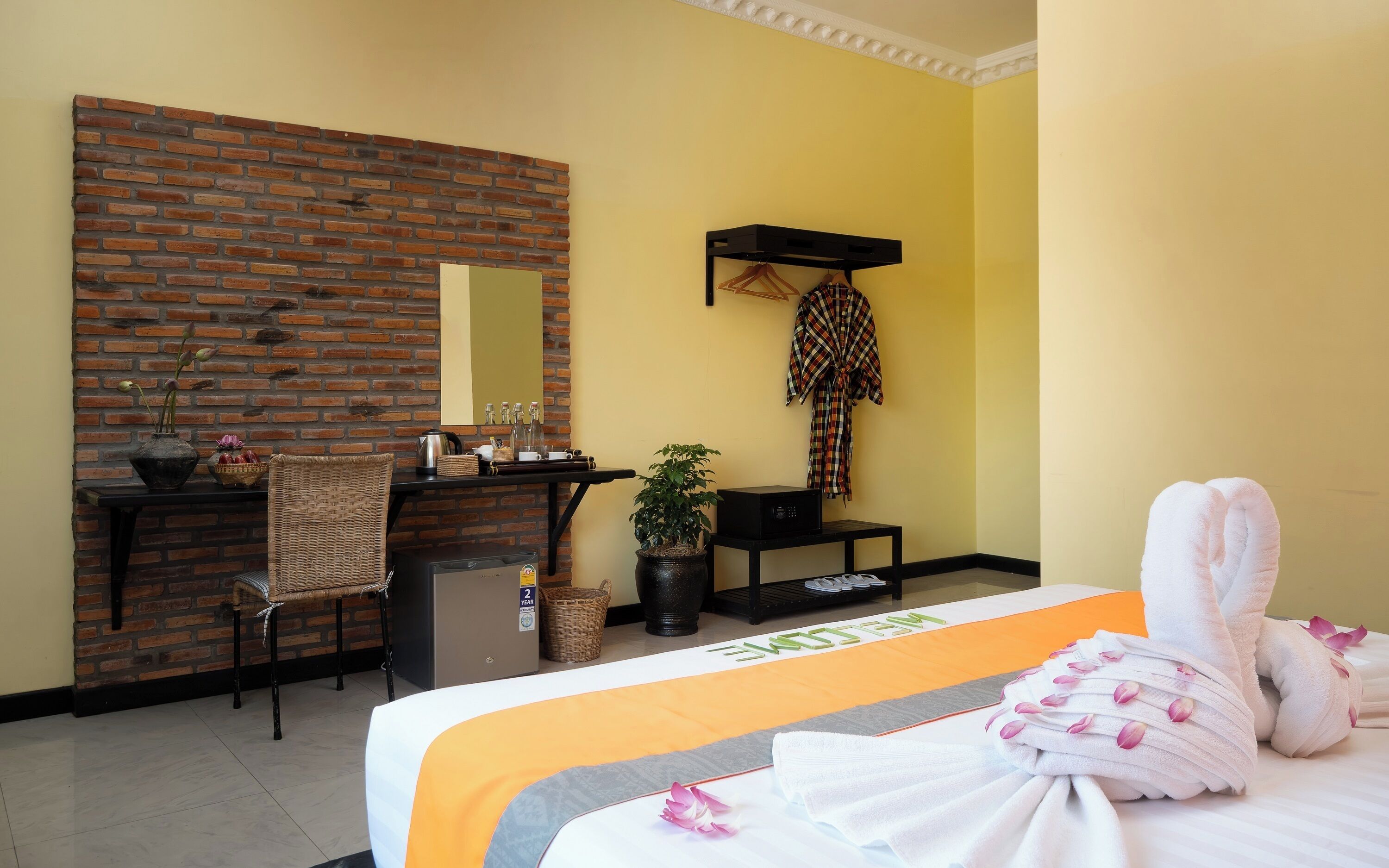 Een hotelkamer met een bakstenen muur, een bureau, een stoel, een bed met handdoeken, een potplant en een spiegel.
