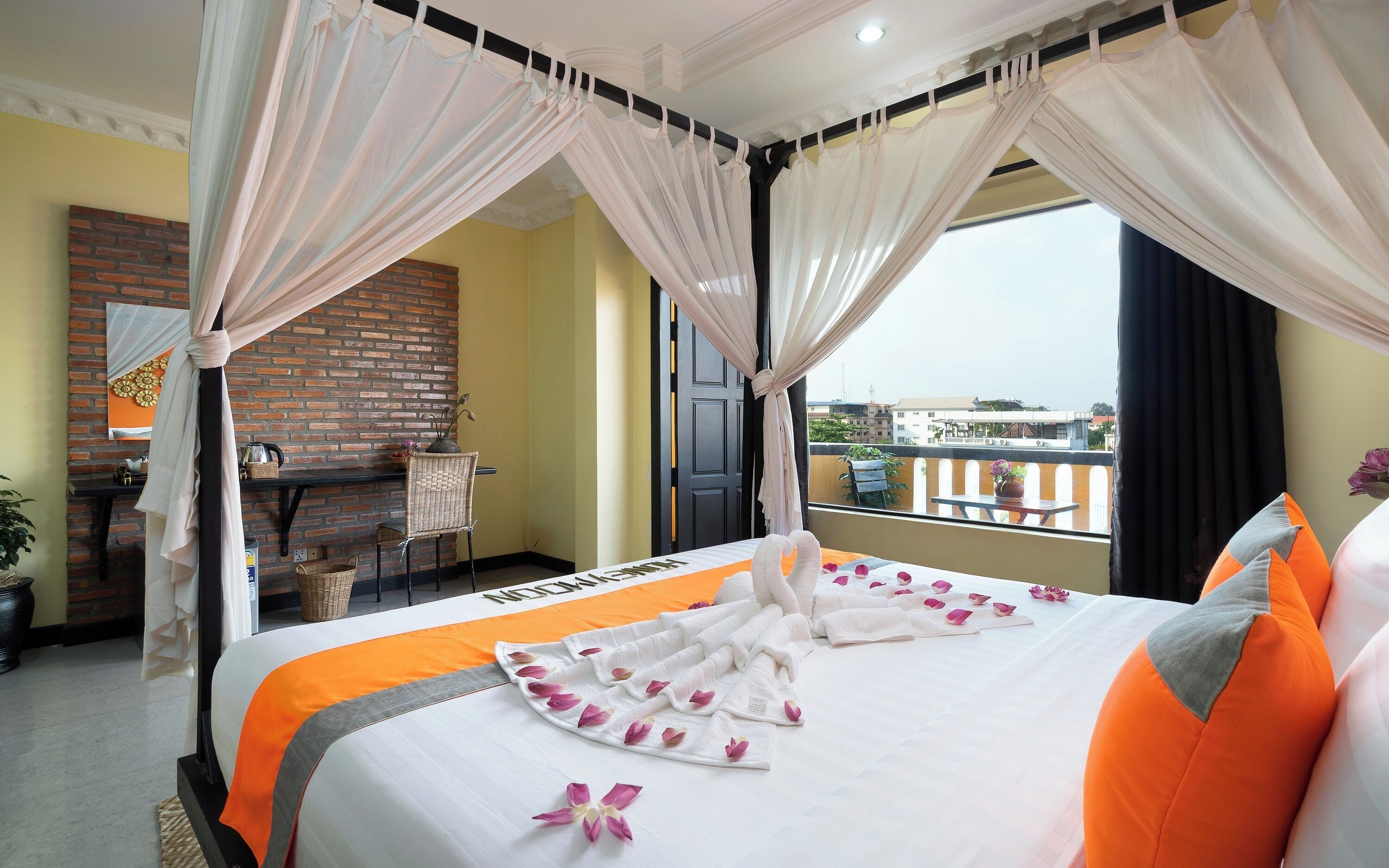 A hotel room with a bed, a small table, a chair, a balcony with a view, and a brick wall.