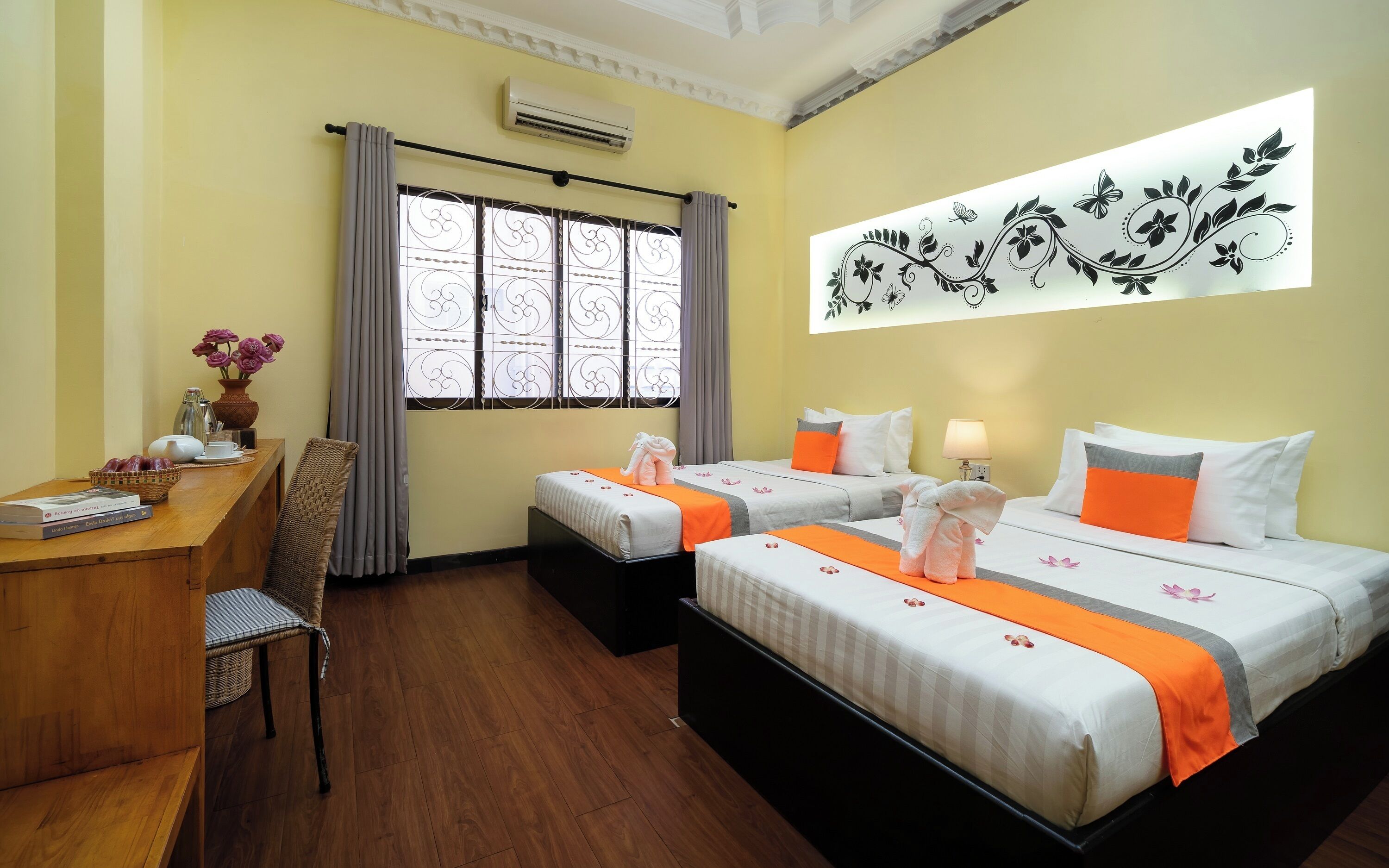 A hotel room with two beds, a desk, a chair, a window with curtains, an air conditioner, and a wall decoration.