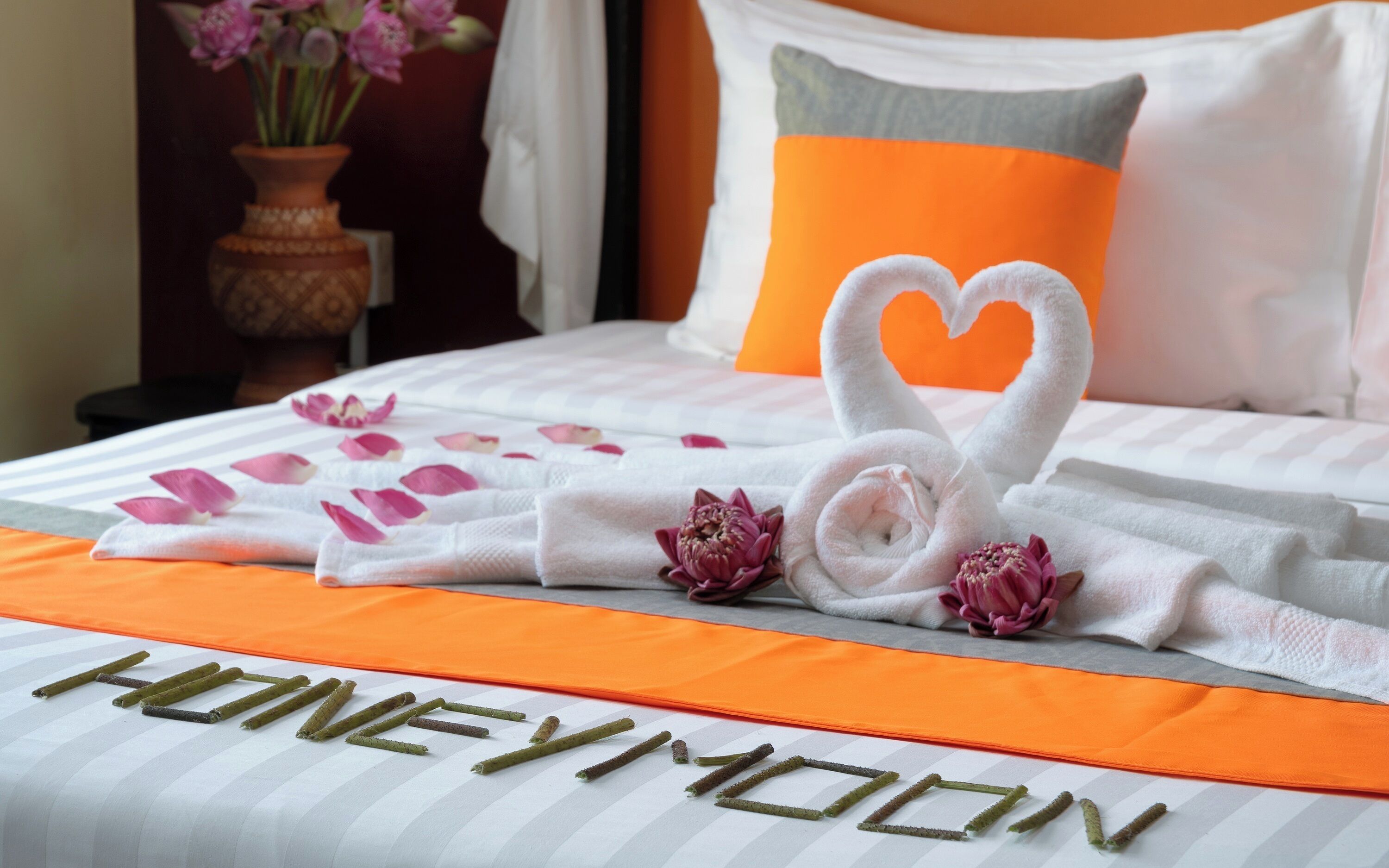 A bed with a white and orange bedspread, a heart-shaped towel arrangement, and a 'honeymoon' sign.