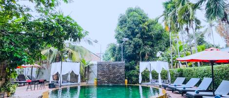 Relax by the serene pool surrounded by lush greenery and palm trees, with lounge chairs and umbrellas for a comfortable stay.