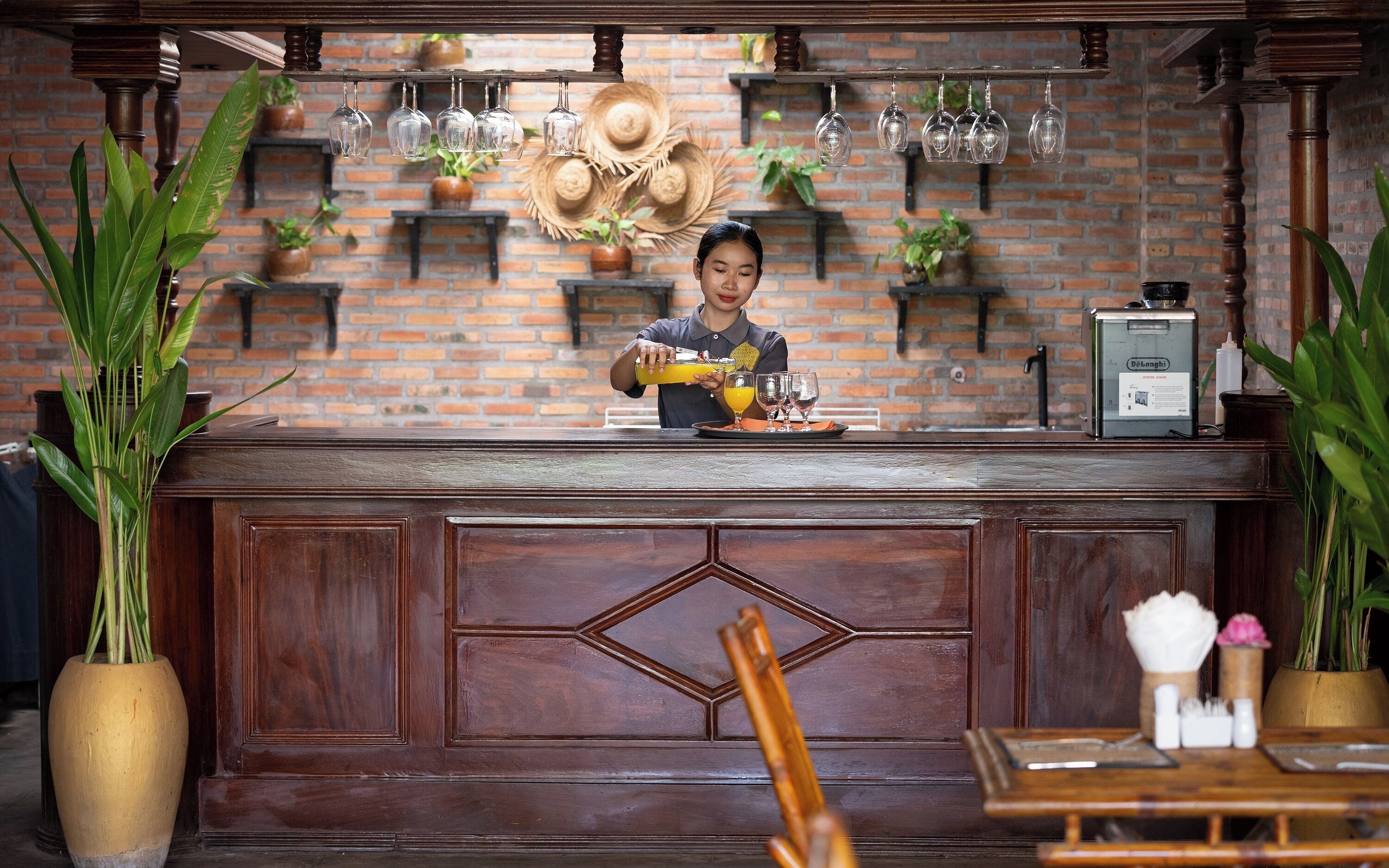 Uma área de bar com parede de tijolos, copos de vinho pendurados e uma pessoa preparando bebidas.