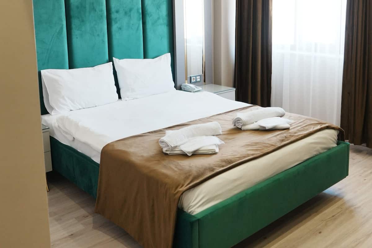 A modern hotel room with a large bed, teal upholstered headboard, white bedding, and a wooden floor.