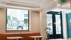 A lobby with a bench, two tables, a potted plant, a view of a street, and a black door with a green exit sign. - Reyyan Hotel (Istanbul)