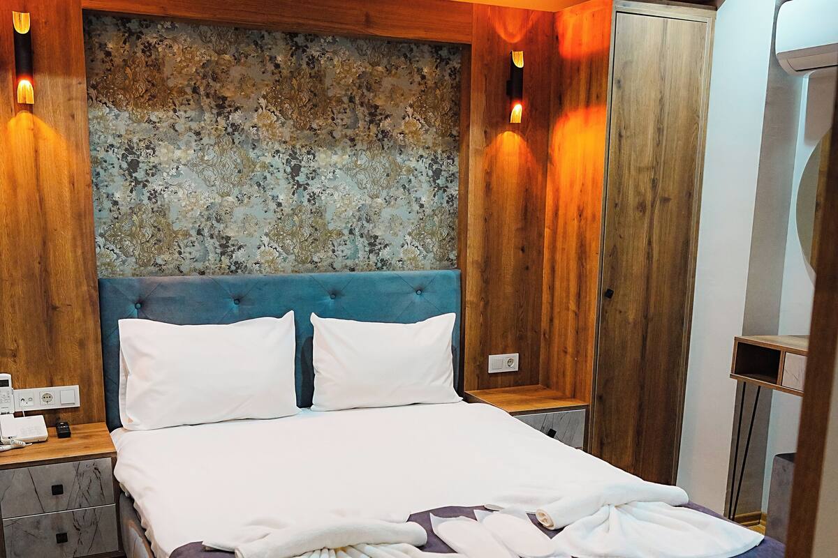 A neatly made bed with white linens and a blue headboard, wooden paneling on the walls, and a small bedside table with a lamp.