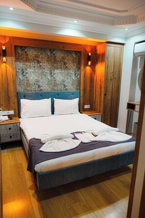 A neatly made bed with white linens and a blue headboard, wooden paneling on the walls, and a small bedside table with a lamp. - Reyyan Hotel (Istanbul)