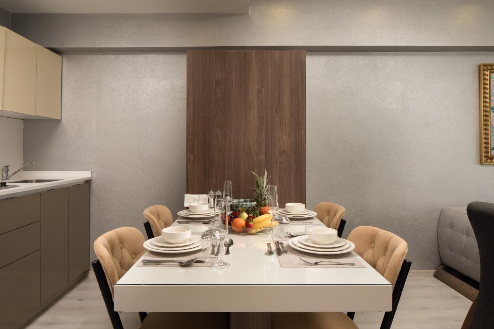 Un'area pranzo con un tavolo bianco apparecchiato per sei persone, circondato da sedie beige, una parete d'accento in legno e un angolo cottura con lavello e pensili.