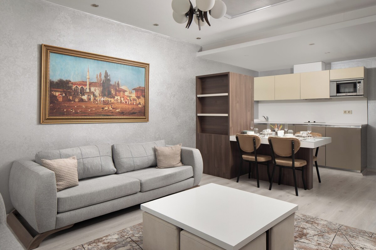 Un salon moderne comprenant un canapé, une table à manger avec des chaises, un coin cuisine avec des placards et un four à micro-ondes, ainsi qu’un tableau encadré accroché au mur.