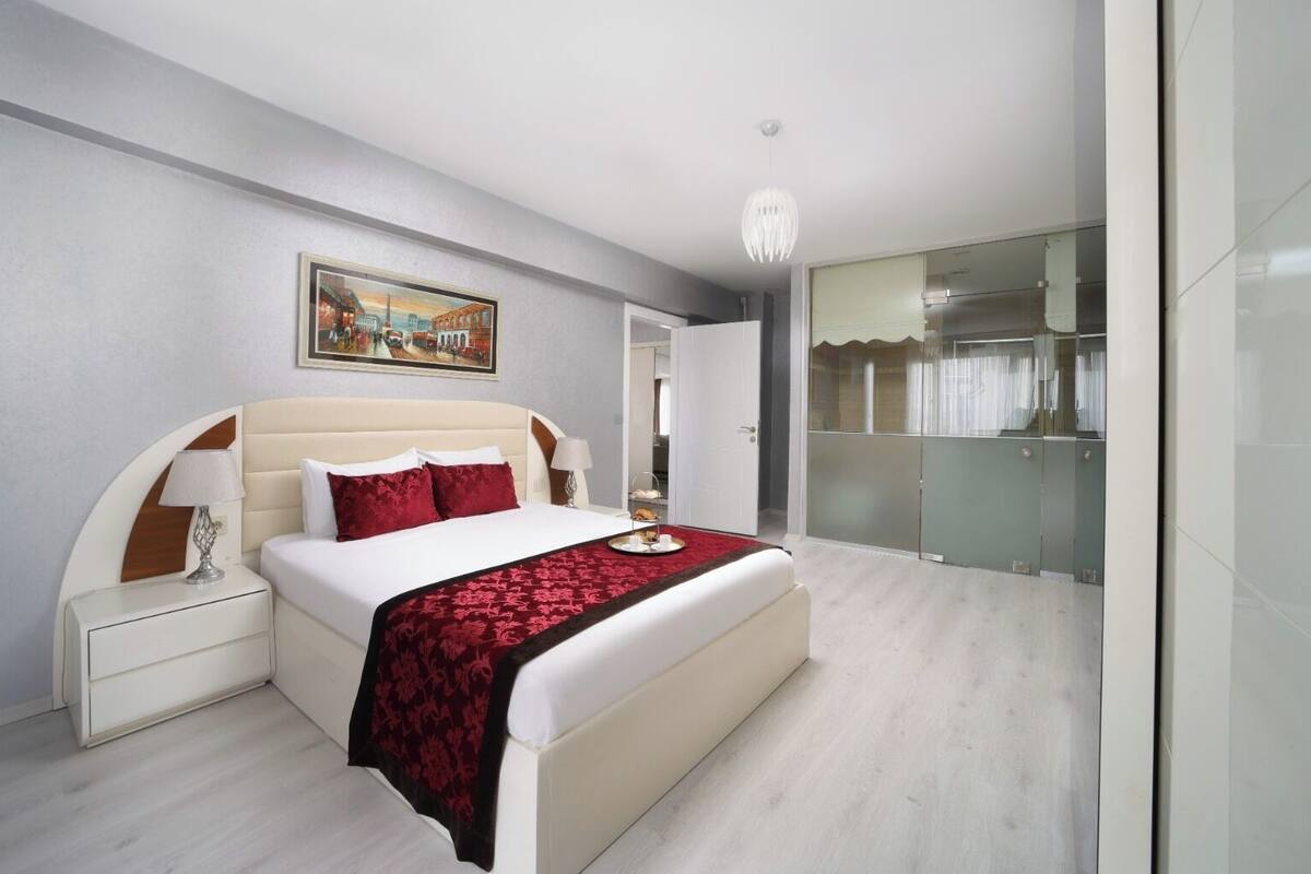 A modern bedroom with a large bed, a bedside table, a nightstand, a painting on the wall, and a glass-enclosed closet.