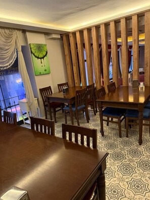 A dining area with a wooden table and chairs, patterned floor tiles, a wall-mounted painting, and a view through a window with curtains.