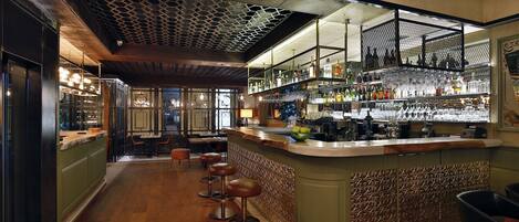Zona de bar con mostrador de madera, taburetes y estantes llenos de botellas y vasos.