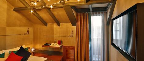 A hotel room with a bed, a TV, a small table with a lamp, and a window with curtains.