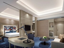 A modern hotel room with a bed, desk, chair, TV, and a small table with flowers.