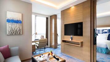 A hotel room with a flat-screen TV, a sofa, a coffee table with snacks, a desk with a lamp, and a bed with a blue bedspread.