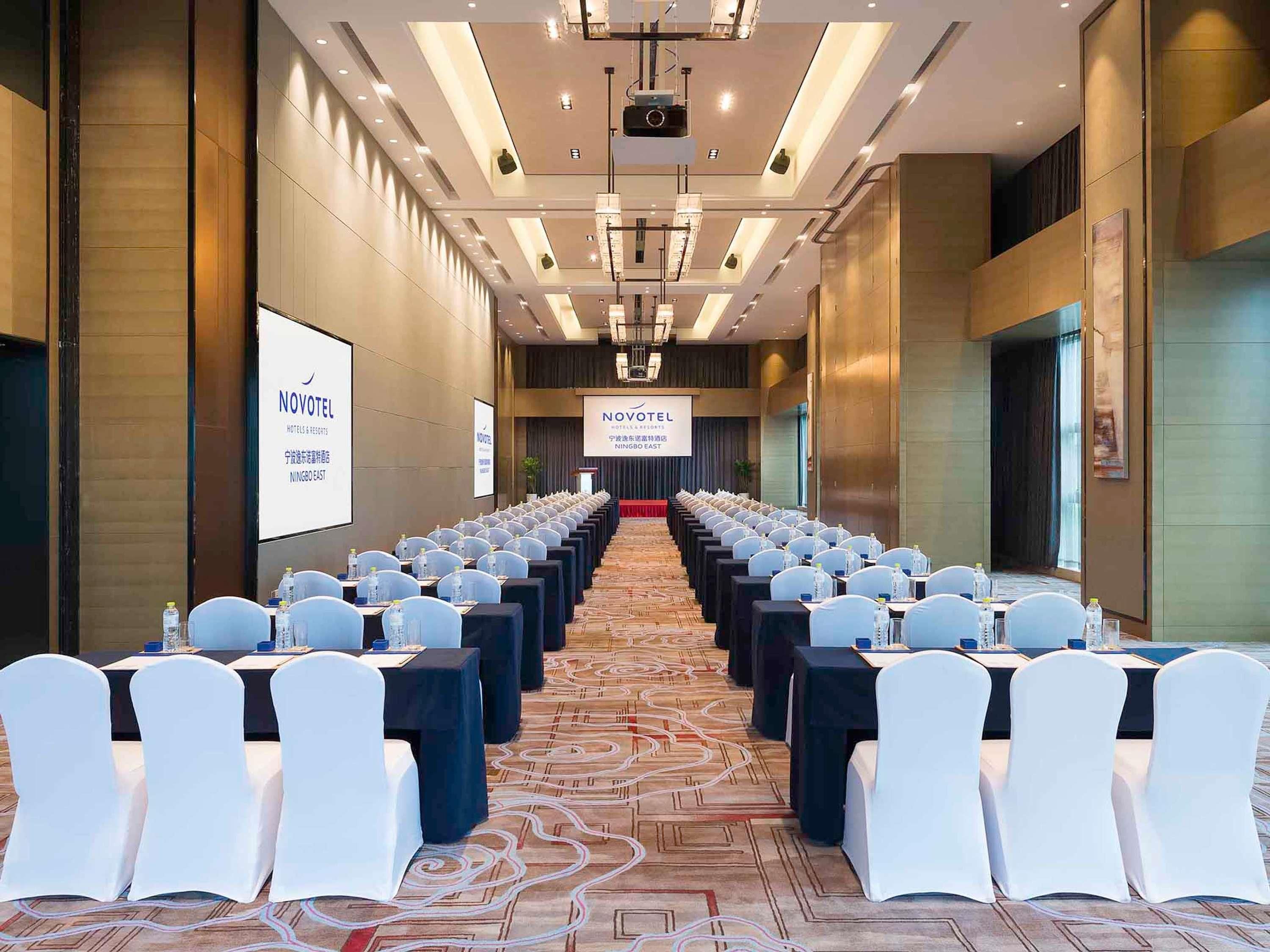 Ein Konferenzraum mit Reihen von Tischen und Stühlen, einer Projektionsleinwand und einem Banner mit 'NOVOTEL'-Branding.