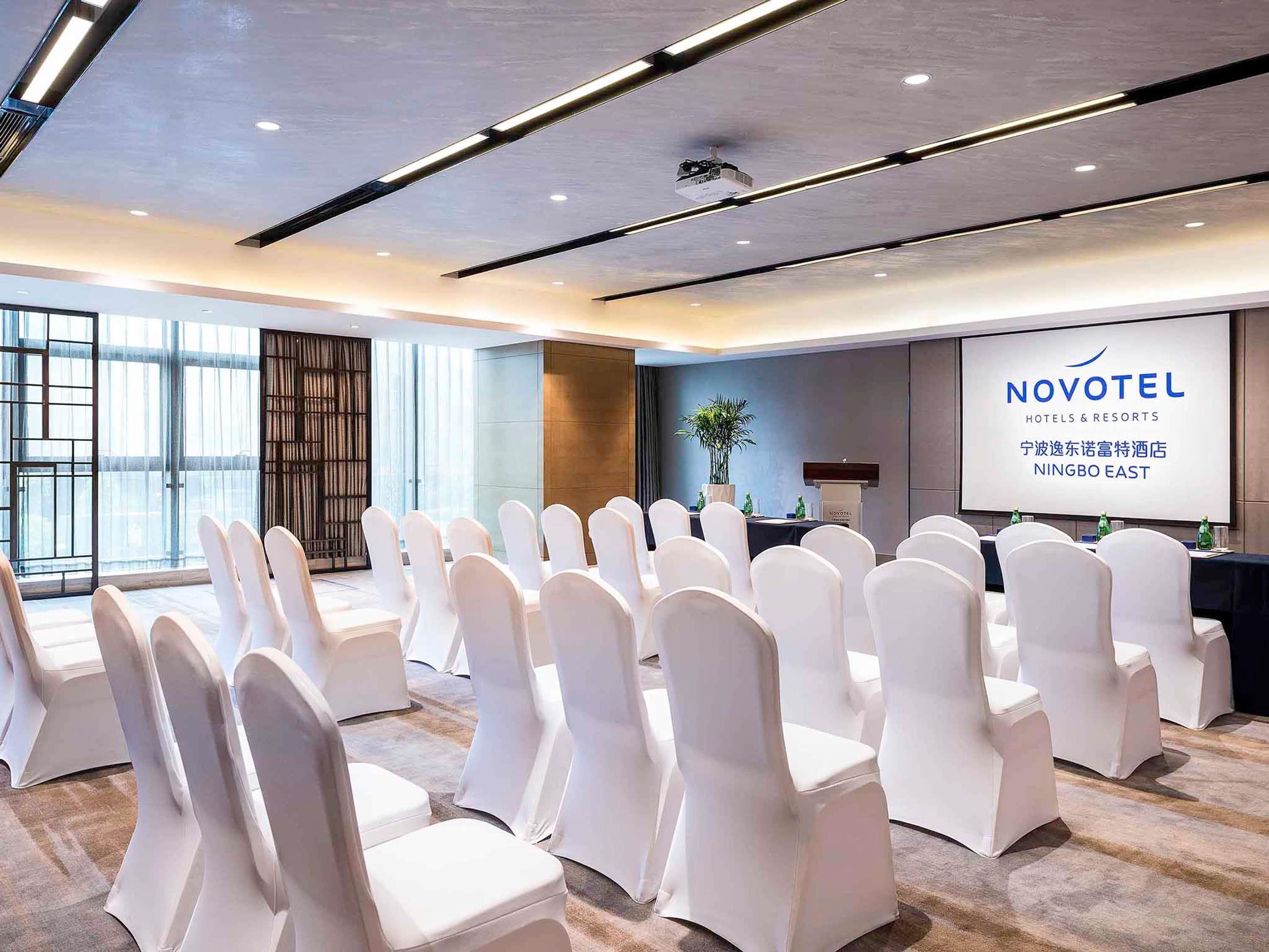 Une salle de conférence avec des rangées de chaises blanches, un pupitre et un écran affichant la mention « NOVOTEL HOTELS & RESORTS NINGBO EAST ».
