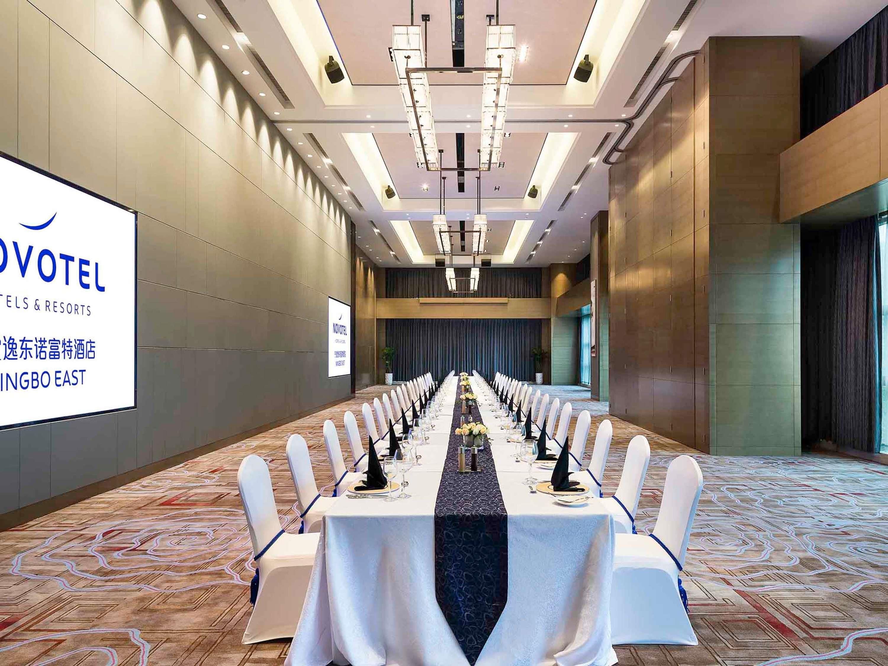 Un salón de conferencias con una larga mesa dispuesta para una reunión, una pantalla grande mostrando texto y un diseño de interiores moderno.