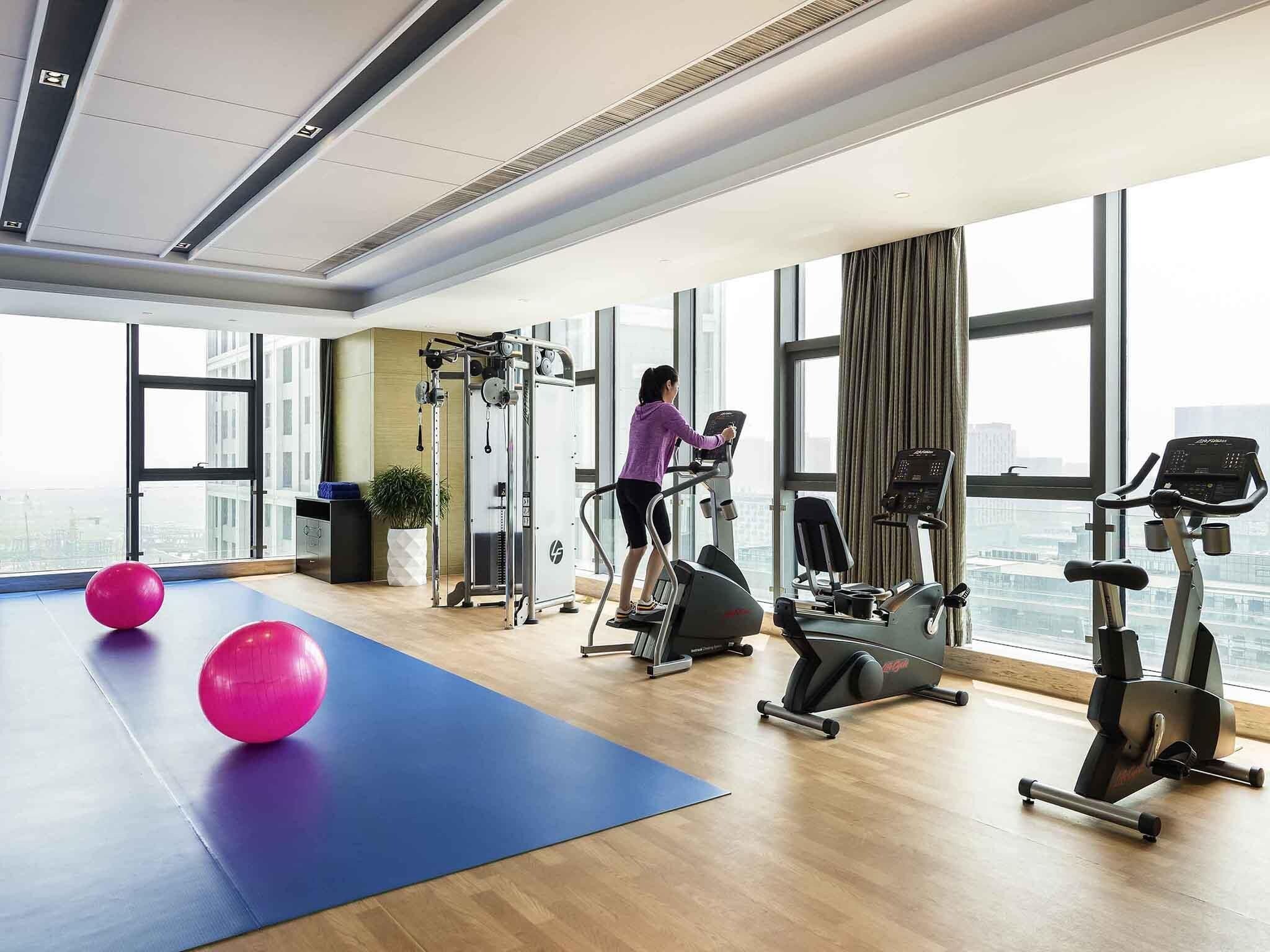 Une salle de sport moderne équipée de machines de cardio, d’un tapis de sol bleu et de ballons d’exercice roses.