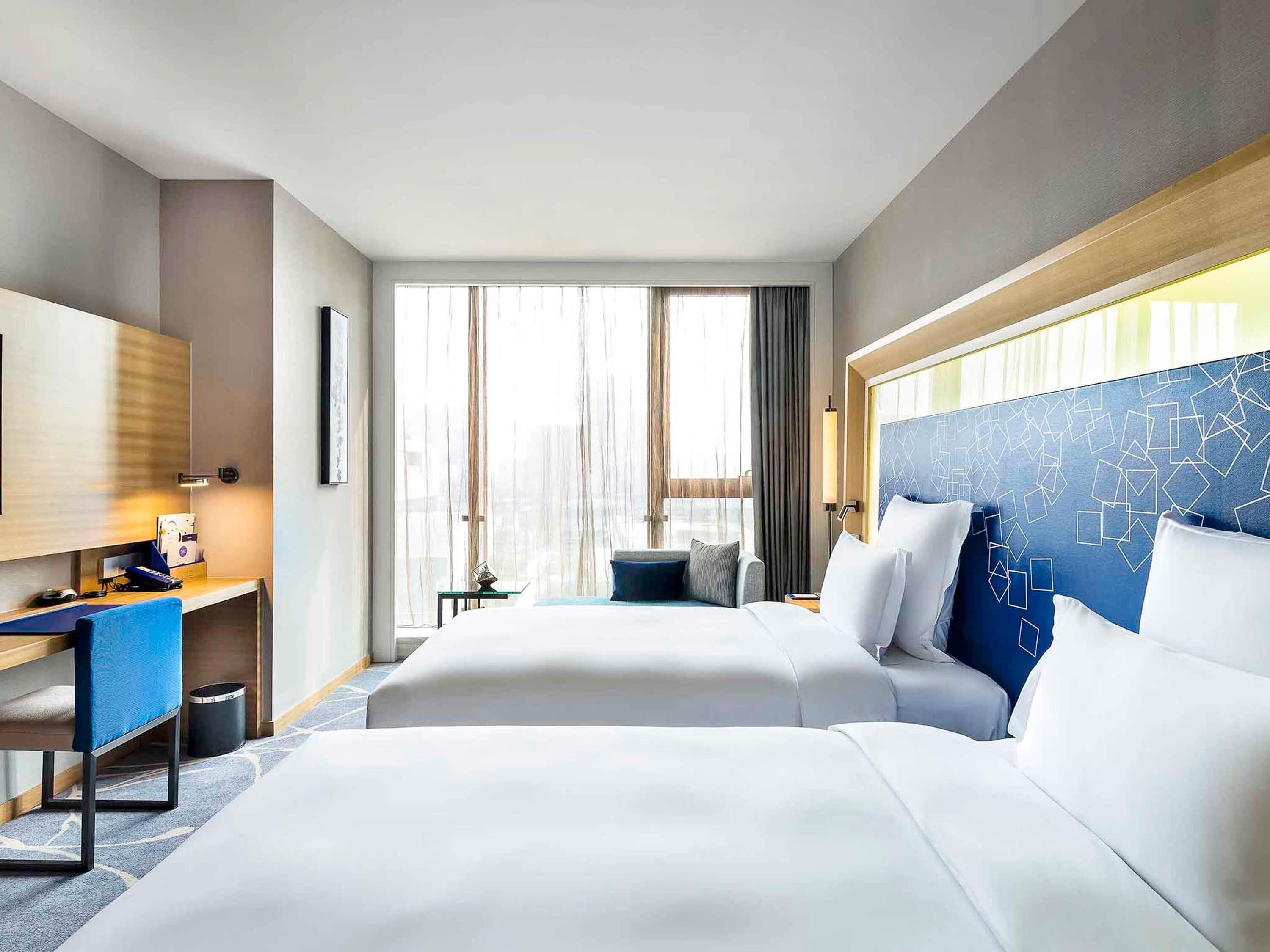 A hotel room with two beds, a desk, a chair, a TV, and a window with curtains.