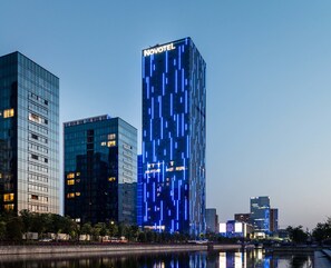 Un hotel moderno de gran altura, con un llamativo letrero 'NOVOTEL', rodeado de otros edificios y una masa de agua.