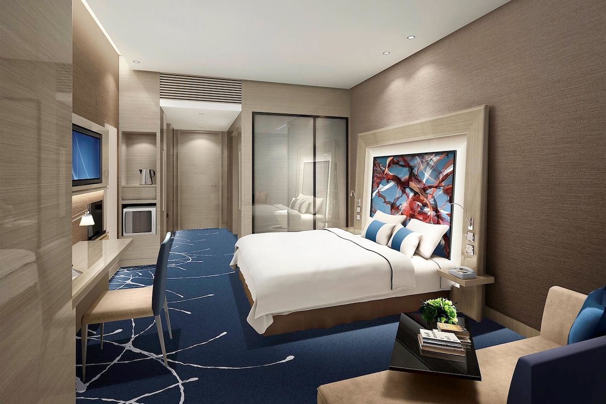 Una habitación de hotel moderna con una cama grande, un escritorio con televisor de pantalla plana, una zona de estar con sofá y sillones, y un cuadro abstracto de gran tamaño en la pared.